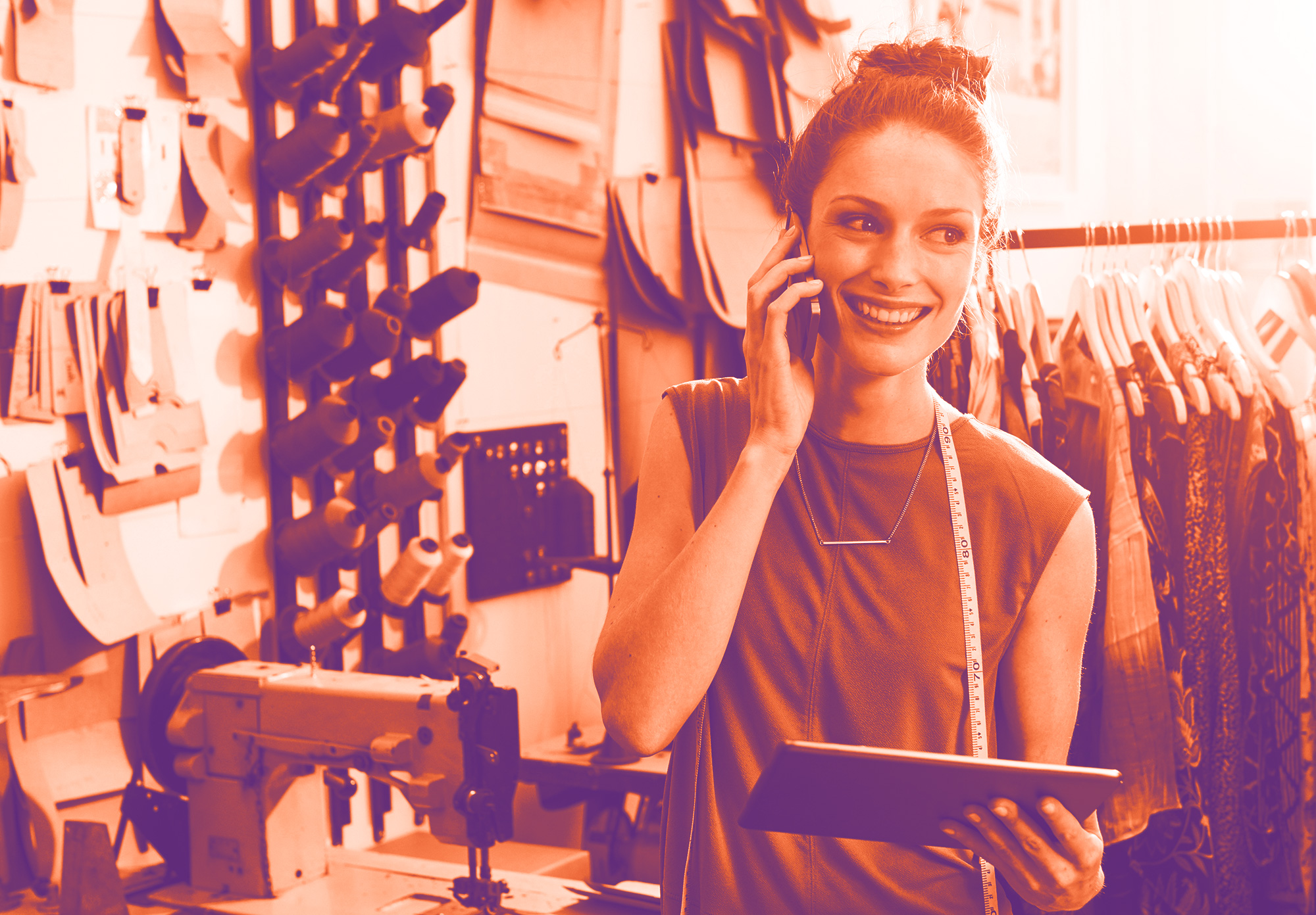 foto con gradiente arancione e viola che rappresenta una sarta all'interno del suo atelier mentre è al telefono e tiene in mano un tablet