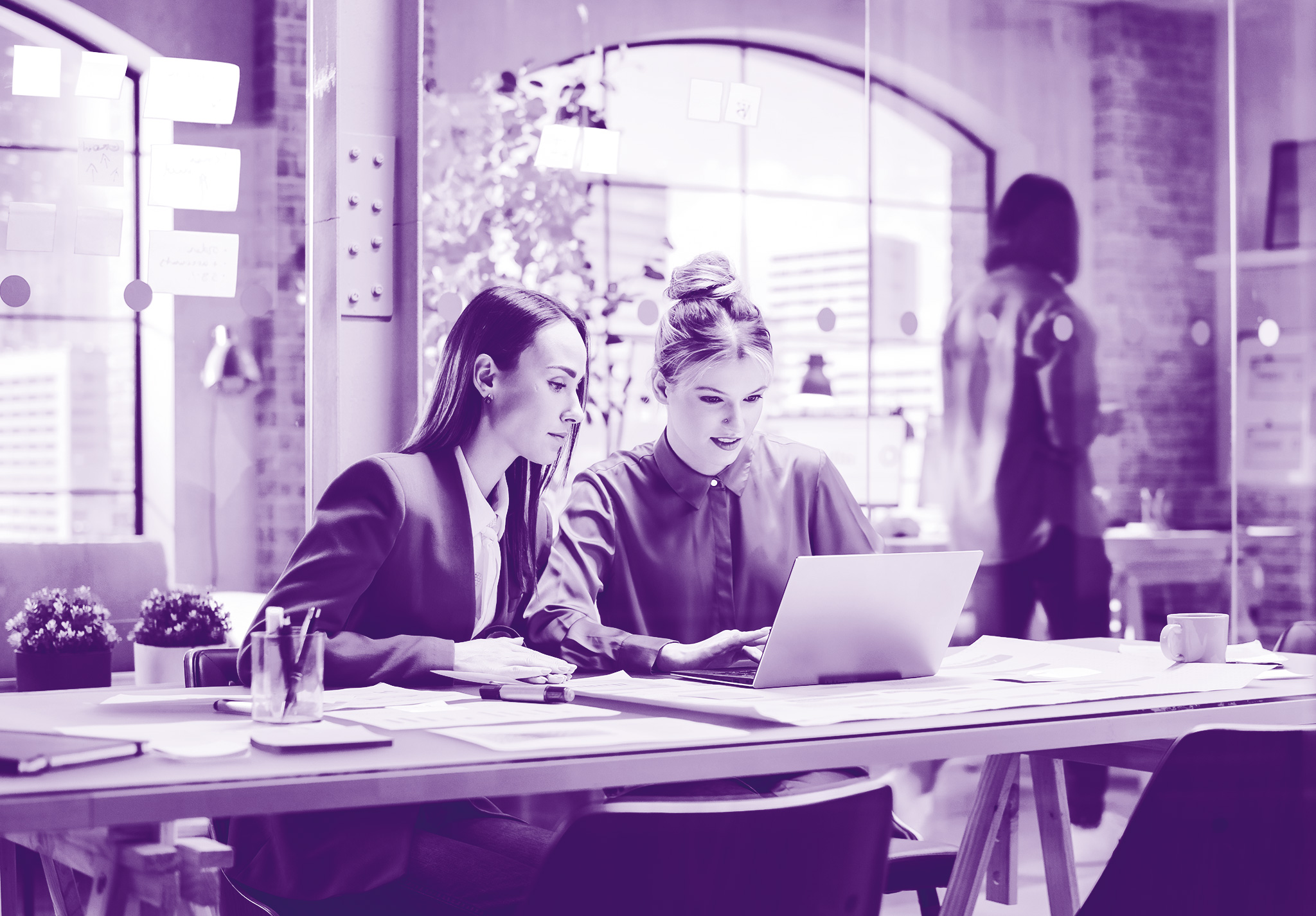 foto con sfondo viola dove ci sono due donne in ufficio sedute alla scrivania che guardano lo stesso pc e si confrontano