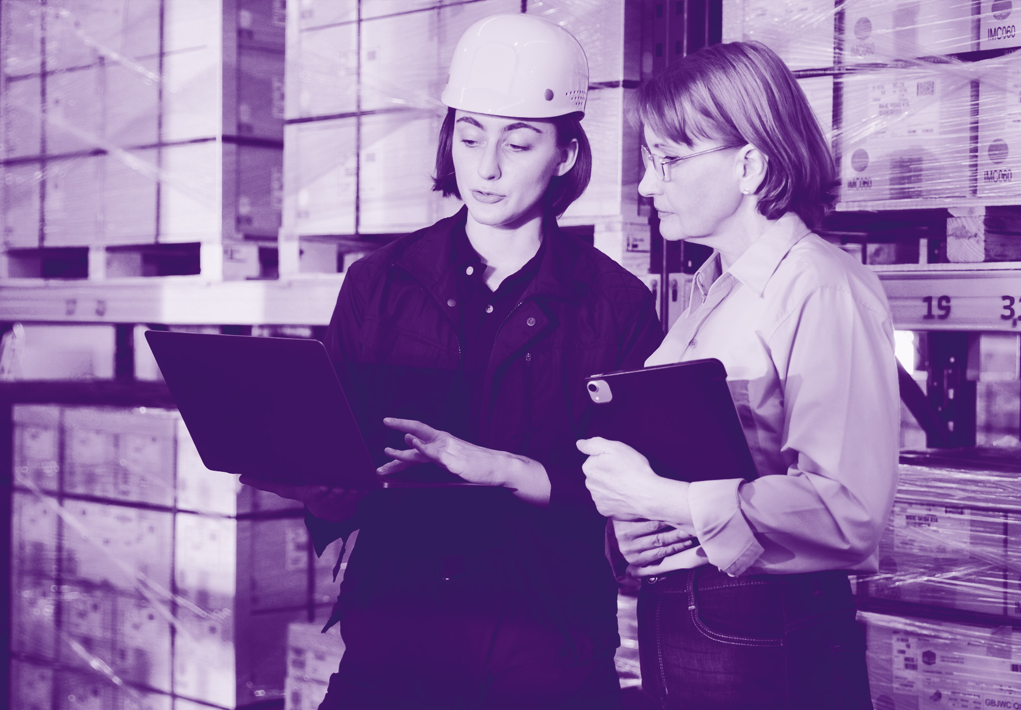 foto con gradiente viola che rappresenta due donne (una con un caschetto da lavoro in testa) che parlano all'interno di un magazzino guardando un pc