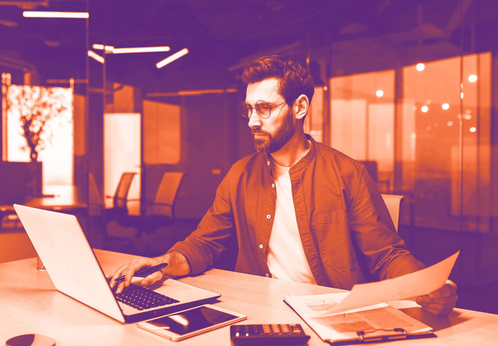 foto con gradiente viola e arancione con un uomo alla scrivania che lavora al computer e con una mano scrive alla tastiera e con l'altra tiene in mano delle carte. Sulla scrivania ci sono anche altri foglia, calcolatrice e tablet