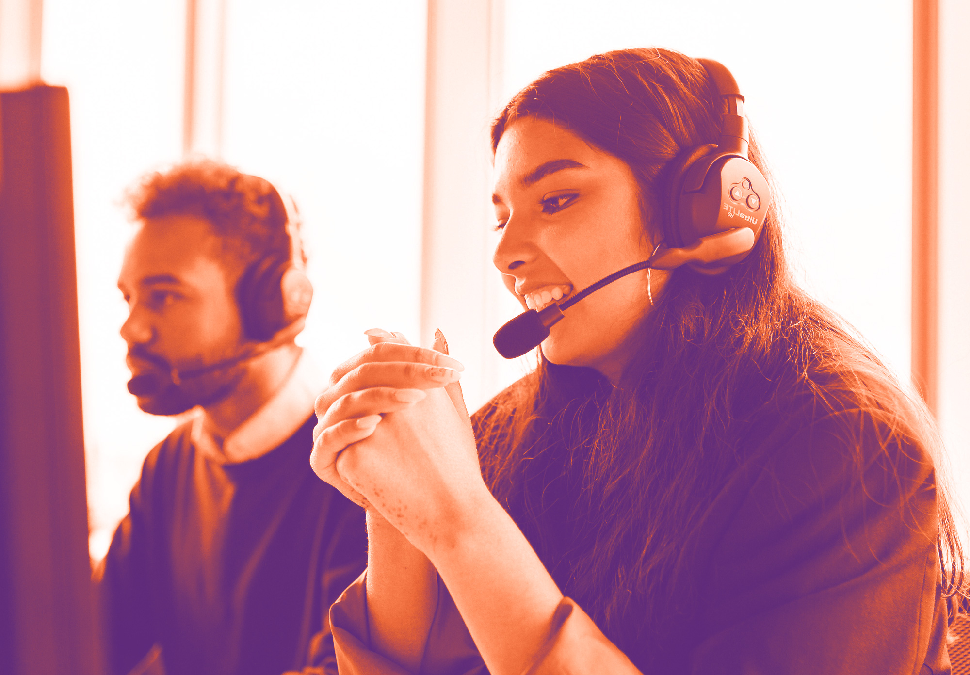 foto con gradiente arancione e viola con una ragazza in primo piano che parla al telefono con le cuffie, sullo sfondo un ragazzo sempre al pc con le cuffie