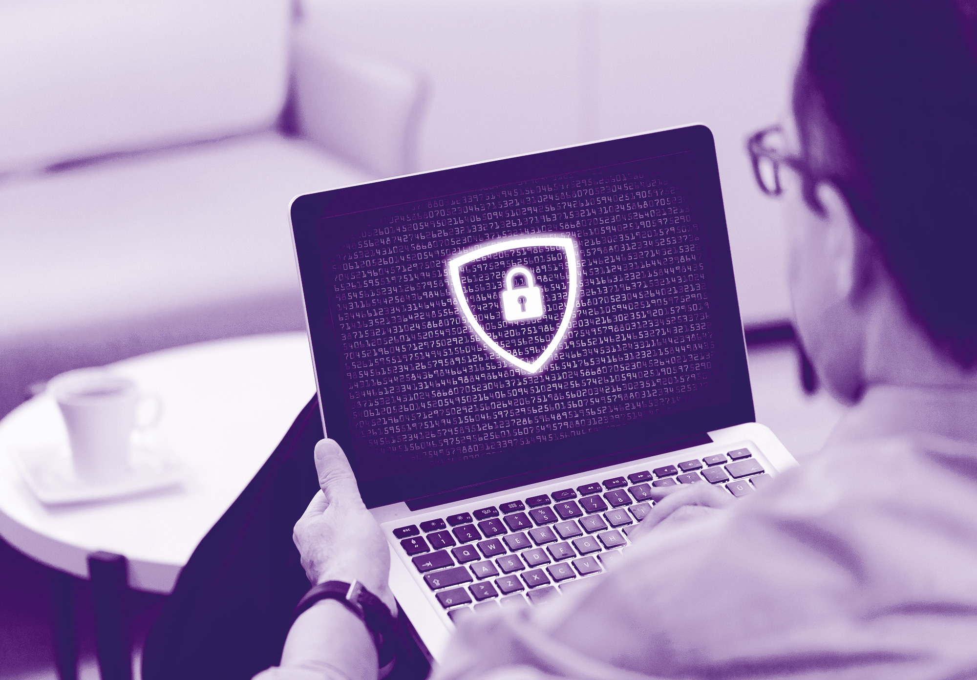 foto con gradiente viola dove c'è una persona di spalle che tiene in mano un pc portatile dove sul monitor si vedono codici di sicurezza e davanti sempre uni scudo con un lucchetto