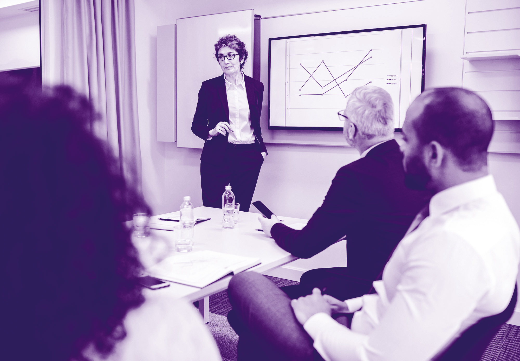 foto con gradiente viola dove si vedono delle persone sedute attorno ad un tavolo e una donna che sta spiegando qualcosa indicando una lavagna appesa al muro che ritrae dei grafici