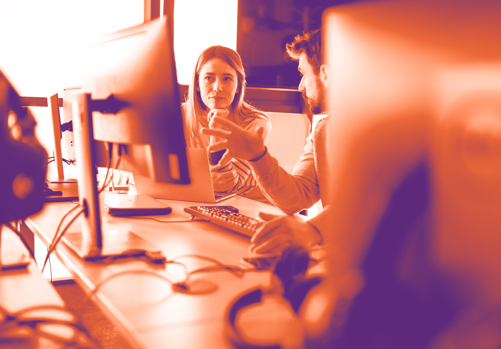 foto con gradiente arancione e viola dove in primo piano ci sono dei Pc sfuocati e poi tra due si possono notare un uomo e una donna seduti alla scrivania che lavorano su un monitor di un pc comune